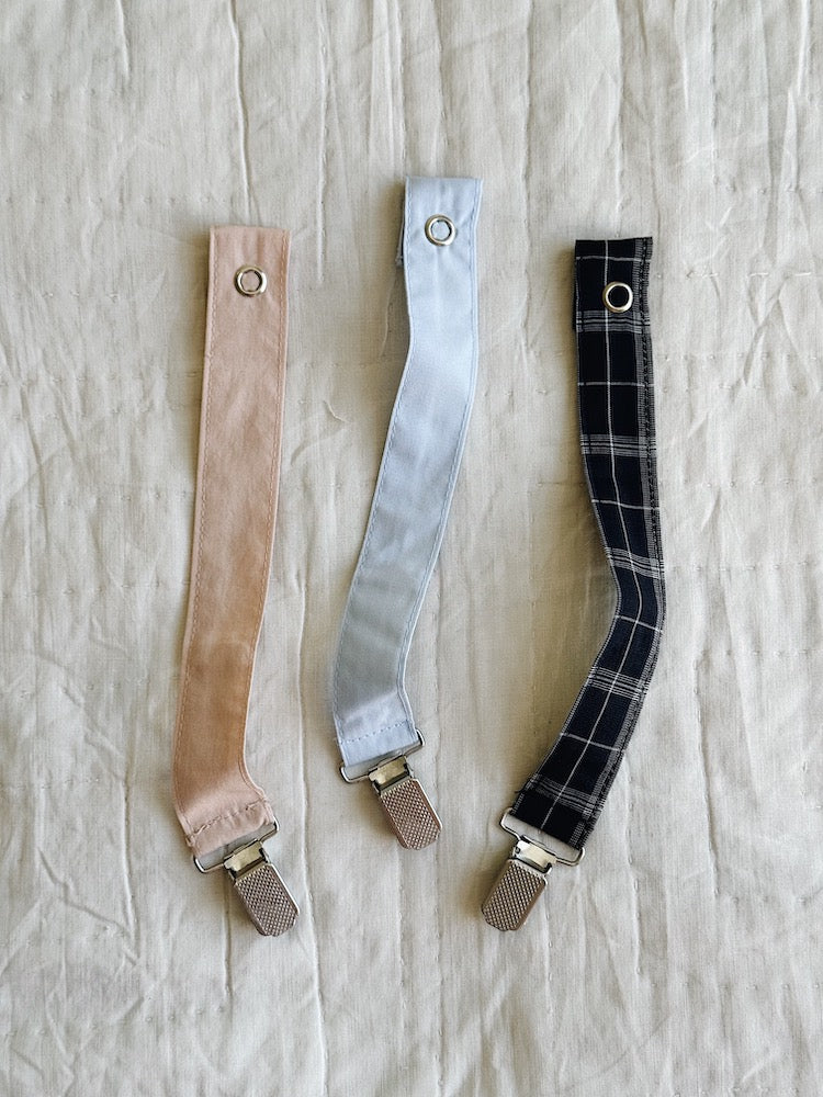 Three pacifier clips for babies, in blush pink, sky blue and blue check, with a metal clip for attachment